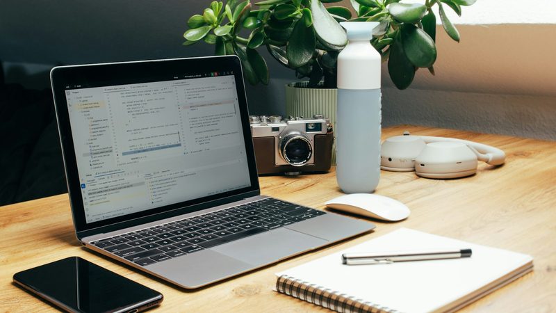 A laptop, phone, vintage film camera and open notebook with a pen on a wooden desk